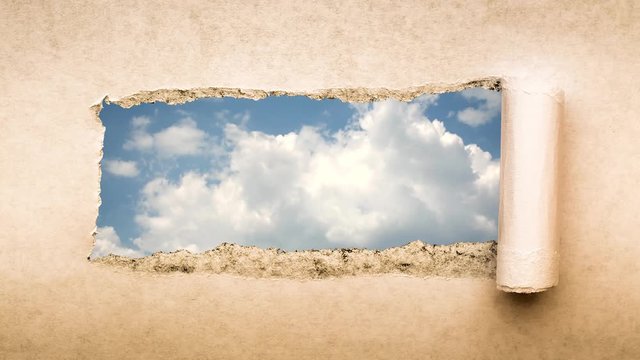 Creative 4k time laps video of fast moving clouds in the blue sky that are visible through a hole with torn edges in old retro grunge vintage paper.