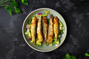 Grilled small fish in a gray plate on a black, dark concrete background. Photo of food. Fried Fish, lettuce salad, parsley, sweet and sour sauce. Top view, place for text.