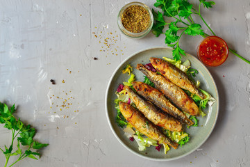 Grilled small fish in a gray plate on a gray concrete background. Food photo, top view. Fried Fish, lettuce salad, parsley, sweet and sour sauce.