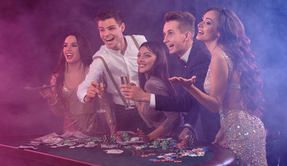 Friends playing poker at casino, at table with stacks of chips, money, cards, champagne on it. Celebrating win, excited. Black background. Close-up.