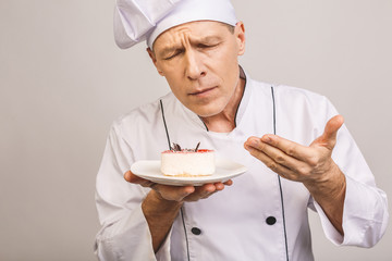 Portrait of a happy senior male chef dressed in uniform holding plate with piece of cake and looking at camera isolated over gray background.