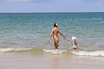 Young mom leading kid to swimming in the sea. Slim woman in bikini with little child on a sandy beach, concept of single mother, family leisure, safety on a water