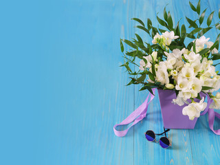 A delicate bouquet of white freesia in a lilac basket and blue glasses on a blue wooden table. Spring card