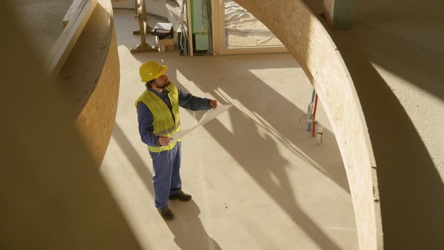 SLOW MOTION: Young Caucasian architect looks at his plans in an unfinished prefabricated house. Static technician in blue workwear and yellow hard hat looks at floor plans and observes the house
