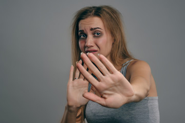 Blonde girl with bruises on her face, trying to protect herself, posing on gray background....