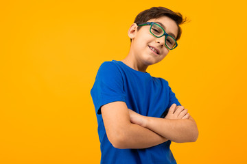 cool teenager boy in a blue tank top and glasses with arms crossed on a yellow background with copy...