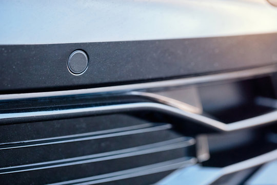 Selective focus on parking sensor of parking assistent system at front of luxurious car bumper close up on light blurred background.