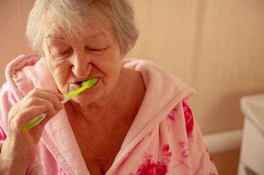 Elderly grandmother in pink terry bathrobe brushes her teeth in bathroom. Hygienic procedures. Pensioners in sanatorium