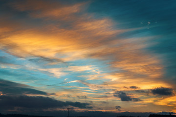 Himmel im Abendlicht mit ziehenden Wolken und Strommast am Horizont in türkis und orange