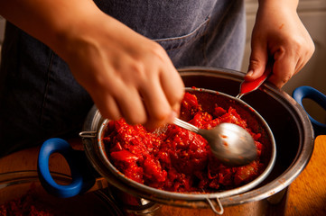 woman cook in an apron prepares tomatoes in a saucepan, rubs through a sieve and prepares tomato juice. Female hands closeup.