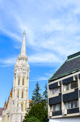 Spire of the Matthias Church in Budapest, Hungary on a vertical photo with adjacent Socialist building. Roman Catholic church built in the Gothic style. Blue sky and white clouds. Eastern Europe