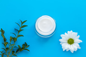 Glass jar of facial cream with white chamomile on blue background, skin care concept. Herbal lotion in bottle, natural cosmetic with daisy.