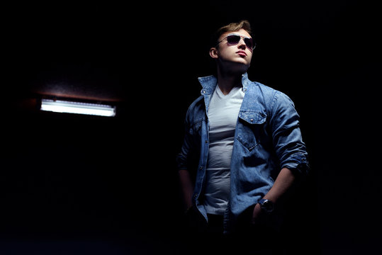 Stylish man posing in light and shadow. Stream of light lit his silhouette. Dressed in sunglasses, denim jacket and white shirt. Bad boy style concept. Dark background.