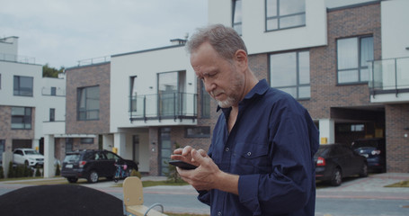 Handsome pensioner using modern mobile gadget walking in the street. Positive mature man in shirt...
