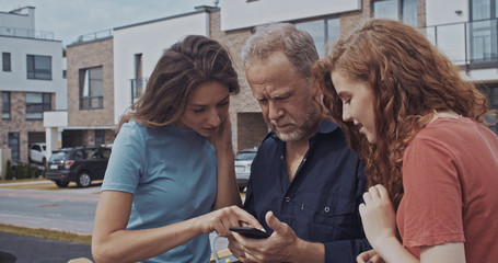 Two positive girls helping older man to deal with online application on smartphone. Polite assistance. Harmony between generations. Modern cityscape background.