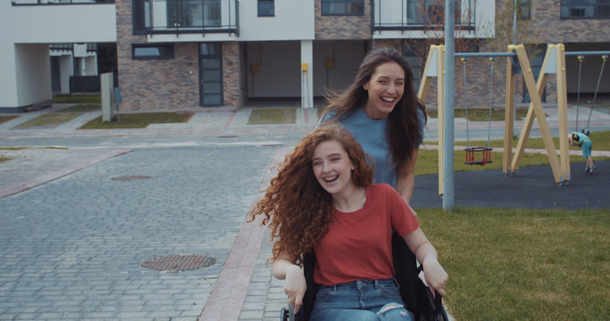 Happy Girl Riding Her Disabled Female Friend In Wheelchair Extreme Fun Sports Activity Leisure Outdoor On Lovely Day In The Modern City. Friendship And Happiness Concept.