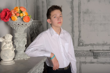 teenager boy in a white shirt and pants leaning on a marble fireplace shelf next to a vase of flowers