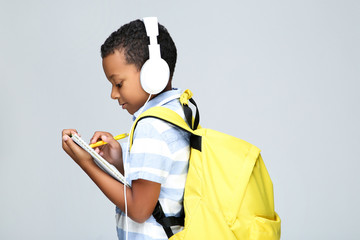 Young African American school boy writing in notepad with headphones and backpack on grey background