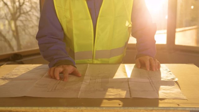 SLOW MOTION, CLOSE UP, LENS FLARE: Golden evening sunbeams shine on male architect in workwear and a hard hat looking at his floor plans. Static technician analyses his plans late in the evening.
