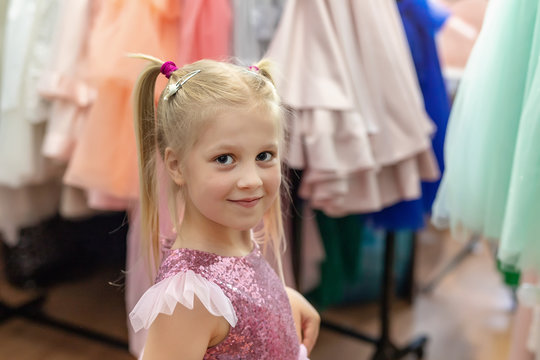 Cute adorable beautiful blond caucasian little girl trying on fashion pink princess dress with sequins at children fashion sewing studio and showroom indoor.Female dress rental salon for event