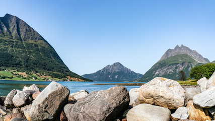 Panoramic view from Luroy at ford with Aldra Island (left), Olvika (center) and some unknown high mountain (left). The wall of heavy stones on sandy beach close photographer, tide time, sunny Norway