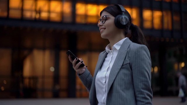 Cheerful spanish young millennial woman enjoying new songs in headphones connected to cellular and online radio having good moot strolling to work