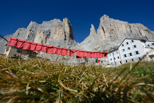 Primo piano della radura erbosa con lo sfondo del rifugio con i panni rossi stesi sullo stendino e delle montagne in contrasto con il cielo blu