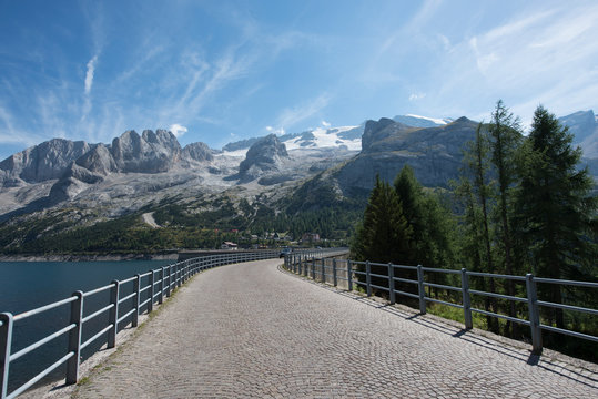 Strada sopra la diga del lago di Fedaia con balaustre ed alberi al lato, sullo sfondo le montagne della marmolada innevate