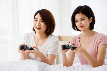 Two Asian girl hold game controller and enjoy video game together on bed. Concept  for teenage or friends activity, lifestyle at home.