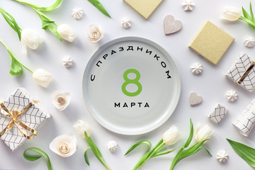 March 8 International Women day celebration. Flat lay, top view on white background with tulip flowers, coffee cups, gifts, sweets. Text in Russian on plate means "with March 8 lovely ladies".