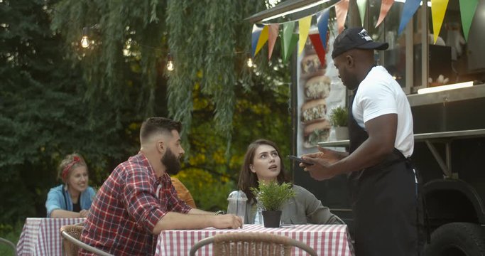 African American young handsome and cheerful male barman in apron and cap asking for order from happy Caucasian couple at table outdoor small food truck cafe in park. Man waiter serving woman and man.