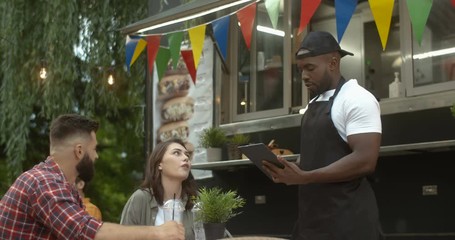 Caucasian happy young couple sitting at table outdoors in park at truck bar. Man and woman ordering food and drinks from African American male waiter with tablet. Bartender from small cafe at work.