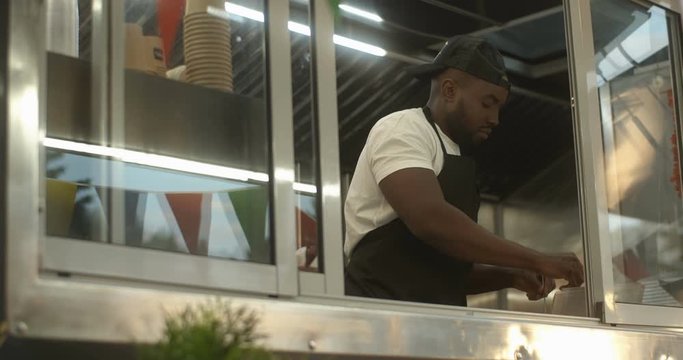Look from outside on festive food truck window with vendor cooking and preparing an order. African American male young bartender making dish at small kitchen of outdoor cafe.