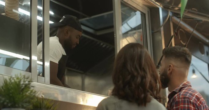 Rear of Caucasian couple standing outdoors at festive food truck cafe window and talking with African American young vendor. Barman of little street bar taking orders from clients.