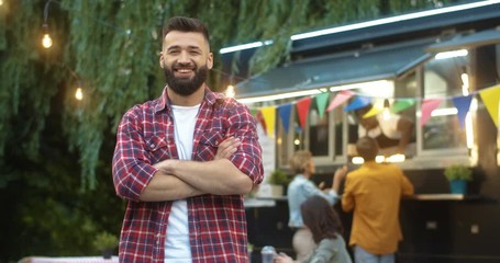 Portrait shot of Caucasian handsome young man standing in front of camera and smiling cheerfully outdoors. Male person looking straight with smile on face. Small festive food truck bar in park.