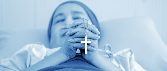 christian cross in hands of cancer patient wearing headscarf during chemotherapy treatment lying on bed in hospital praying and worship  for hope, in monotone