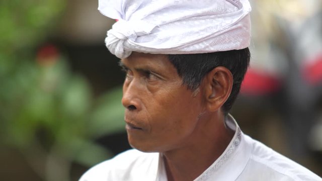 Brown skinned Balinese man in white clothing exhales smoke through his nose then raises cigarette to lips and inhales.  Man in Bali smoking outside in tropical garden.
