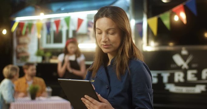 Caucasian middle-aged attractive woman standing outdoor food truck with tablet device. Beautiful female client of middel age tapping and scrolling on computer in festive food track and smiling.