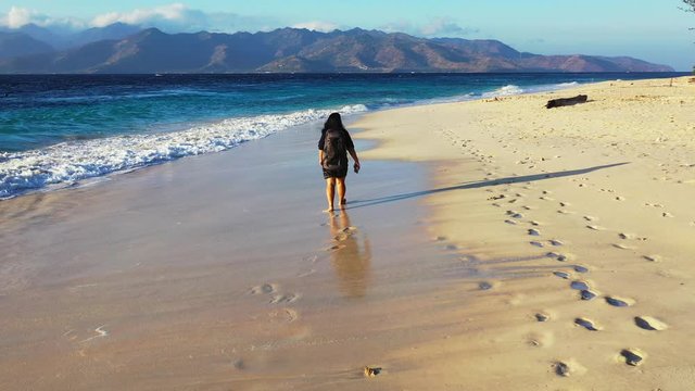 Bali, Indonesia. Woman walking on a long sandy beach with the backpack. Waves fashing the footprints. Mountains in the background.