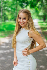 Fototapeta premium Portrait of a charming blond woman wearing beautiful white dress standing on the road under trees.