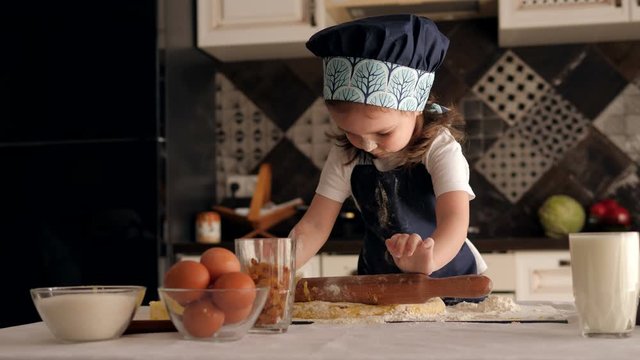 A cute little girl of three years old in a cook's cap and apron rolls a rolling pin dough at home. The child learns to cook homemade cookies. A little girl plays cooks at home in the kitchen.