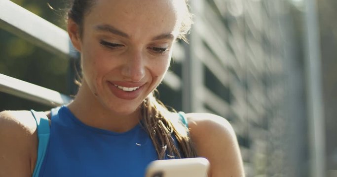 Pretty smiled Caucasian woman in sporty style typing and tapping on smartphone on summer day. Beautiful joyful young girl texting mesage on phone as chatting at sport court.