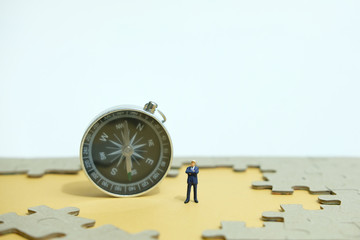 Business strategy conceptual photo - Miniature businessman standing beside compass in the middle of jigsaw puzzle 