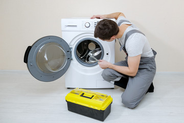 young serviceman in grey uniform repairs broken washing machine with wrench on wooden floor near...