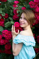 Spring. Close up portrait happy attractive redhead young woman wearing stylish blue light dress smelling blooming roses in the garden. Outdoor.