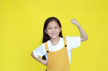 Smiling asian little girl showing his muscle with looking camera isolated on yellow background. Strong kid in dungarees with good health concept