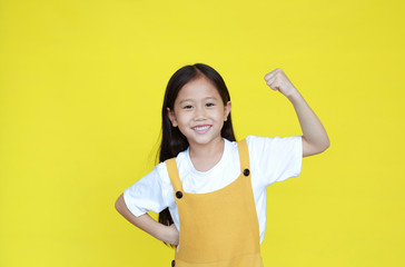 Smiling asian little girl showing his muscle with looking camera isolated on yellow background....