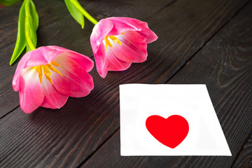 Two fresh pink tulip flowers and white sheet of paper with heart on top lie on dark wooden background. Happy holiday, womens day or wedding greetings concept. Copy space for text.