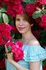 Spring. Close up portrait of a happy pretty redhead girl with charming smile wearing trendy blue light dress on a background of blooming roses. Outdoor.