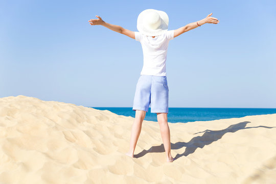 Summer sea vacation. Happy young female arms apart wearing a white hat with a wide brim enjoying life on the sandy beach by the sea on clear sunny day. Back view